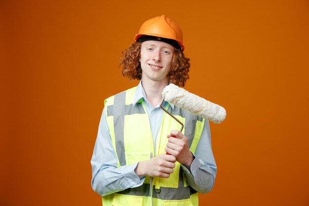 Builder young man in construction uniform and safety helmet holding paint roller looking at camera smiling cheerfully standing over orange background