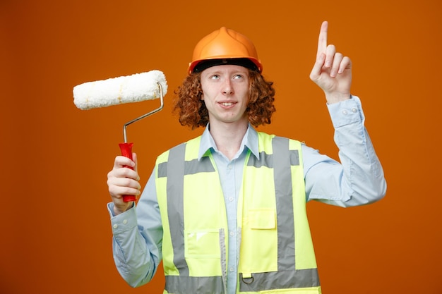 Giovane costruttore in uniforme da costruzione e casco di sicurezza che tiene il rullo di vernice che guarda in alto puntando con il dito indice in piedi su sfondo arancione