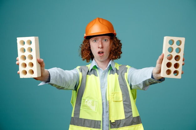 Builder young man in construction uniform and safety helmet holding two bricks looking at camera being worried standing over blue background