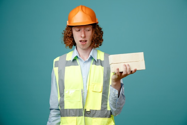 Builder young man in construction uniform and safety helmet holding two bricks looking at them intrigued standing over blue background