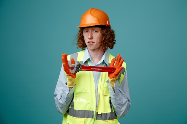 Giovane costruttore in uniforme da costruzione e chiave inglese che tiene il casco di sicurezza guardando la fotocamera con un'espressione seria e sicura in piedi su sfondo blu