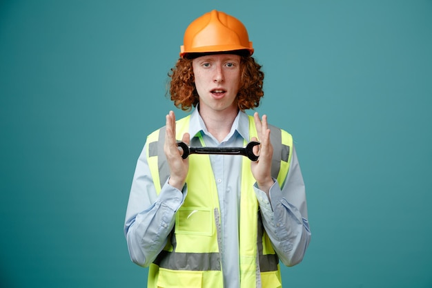 Builder young man in construction uniform and safety helmet holding wrench looking at camera with serious confident expression standing over blue background