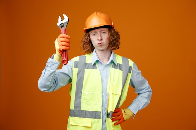 Builder young man in construction uniform and safety helmet holding wrench looking at camera with serious face standing over orange background