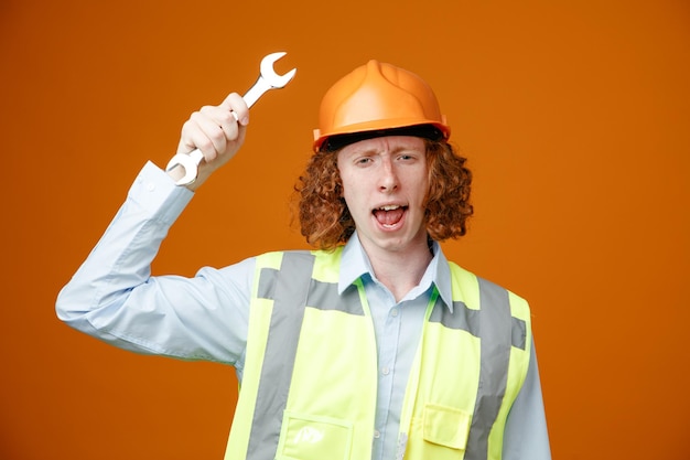 Builder young man in construction uniform and safety helmet holding wrench swinging looking at camera with angry face standing over orange background