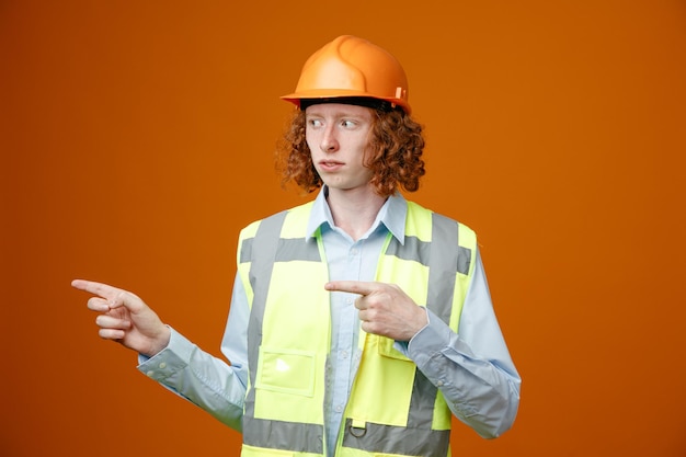 Builder young man in construction uniform and safety helmet looking aside with serious face pointing with index fingers to the side standing over orange background