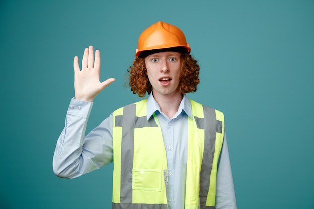 Builder young man in construction uniform and safety helmet looking at camera being surprised waving with hand greeting standing over blue background