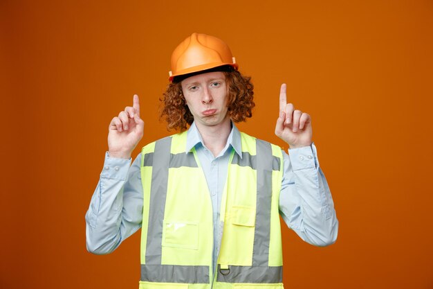 Builder young man in construction uniform and safety helmet looking at camera making wry mouth with sad expression pointing with index fingers up standing over orange background