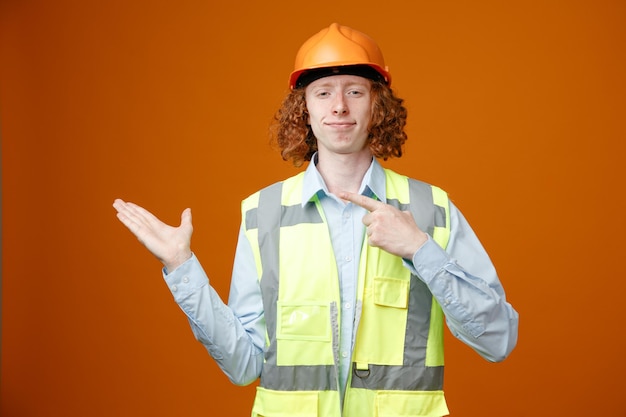 Photo builder young man in construction uniform and safety helmet looking at camera smiling presenting with arm of hand pointing with index finger to the side standing over orange background