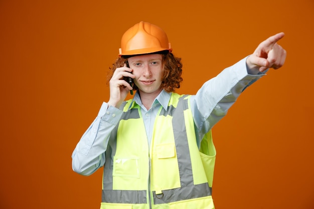 Builder young man in construction uniform and safety helmet talking on mobile phone smiling looking at camera pointing with index finger to the side standing over orange background