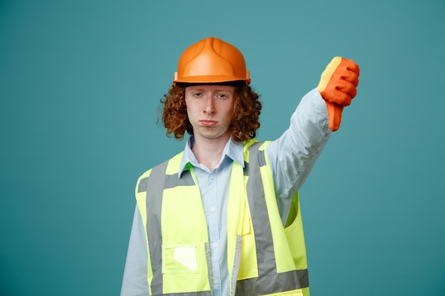 Foto giovane costruttore in uniforme da costruzione e casco di sicurezza che indossa guanti di gomma che sembra insoddisfatto che mostra il pollice verso il basso in piedi su sfondo blu