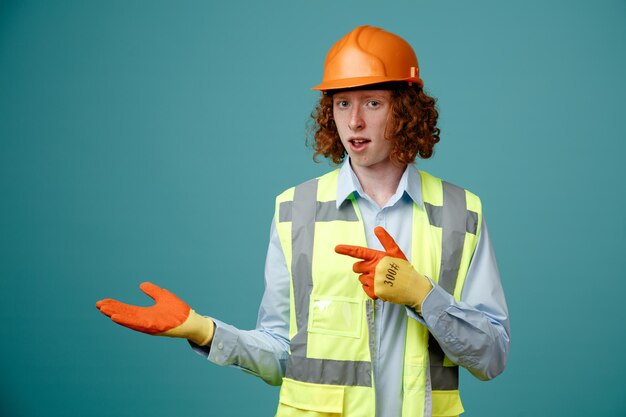 Foto giovane costruttore in uniforme da costruzione e casco di sicurezza che indossa guanti di gomma che presentano qualcosa con il braccio che punta con il dito indice di lato guardando fiducioso in piedi su sfondo blu