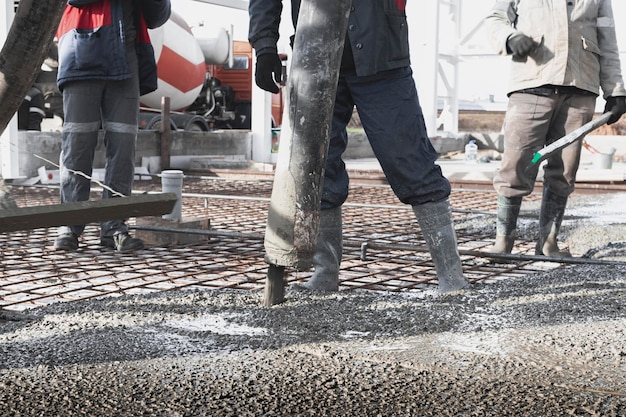 I lavoratori dei costruttori versano il pavimento in cemento nell'officina industriale gambe negli stivali in cemento presentazione del cemento per il getto del pavimento opere monolitiche in cemento