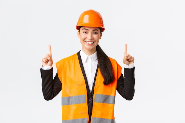 Building, construction and industrial concept. Happy smiling female asian engineer in safety helmet and reflective clothing, introduce new object, estate for sale. Architect pointing fingers up