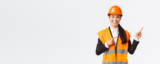 Building construction and industrial concept Smiling female asian engineer in safety helmet and reflective clothing showing object pointing fingers right saleswoman introduce estate to client