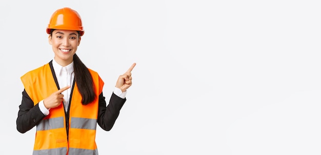 Building construction and industrial concept Smiling female asian engineer in safety helmet and reflective clothing showing object pointing fingers right saleswoman introduce estate to client