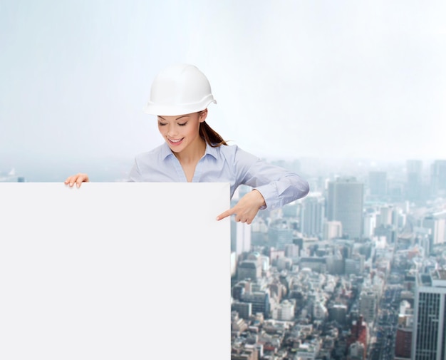 building, developing, consrtuction and architecture concept - young smiling businesswoman in helmet pointing finger to white blank board