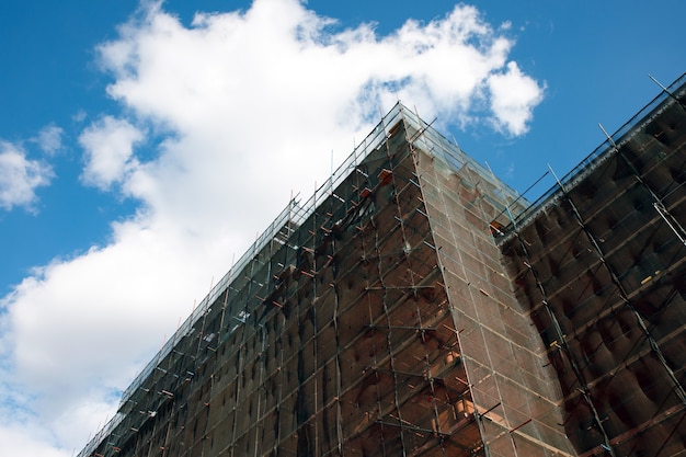 Photo building facade renovation over blue sky, old house reconstruction, repair. scaffold in front of building facade covered with transparent fabric