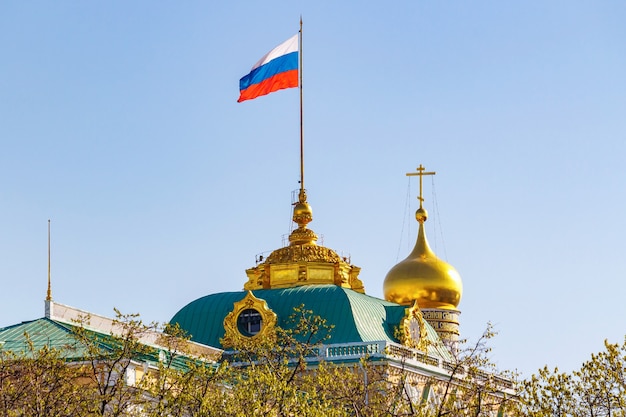 Building of Grand Kremlin Palace with waving Russian Federation flag on the roof against golden dome of church in sunny morning