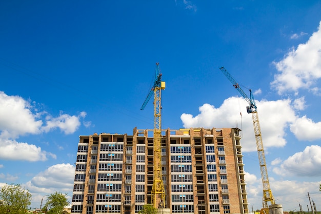 Building site with two industrial tower cranes working at construction of new brick tall building on bright blue sky and green trees scene.