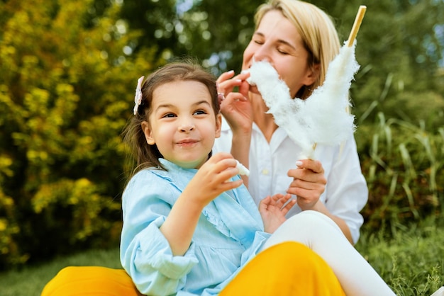 Buiten beeld van gelukkige moeder suikerspin eten met haar dochter zittend op het gras in het park jonge vrouw genieten van de tijd samen met haar schattige kind buitenshuis Moederdag