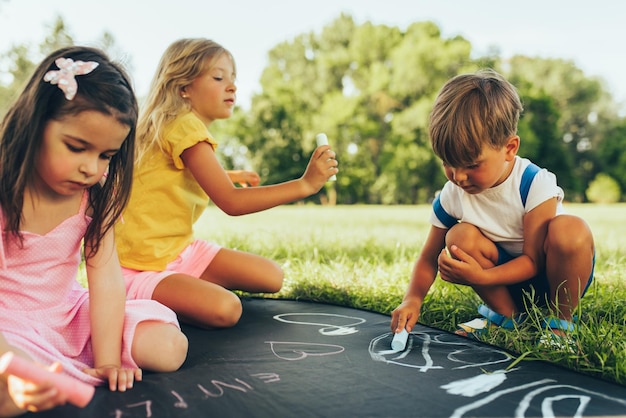 Buiten beeld van kinderen zitten op het groene gras spelen met kleurrijke krijtjes Gelukkige kinderen tekenen met krijt in het park Vrienden jongen en twee meisjes plezier op zonlicht buiten jeugd