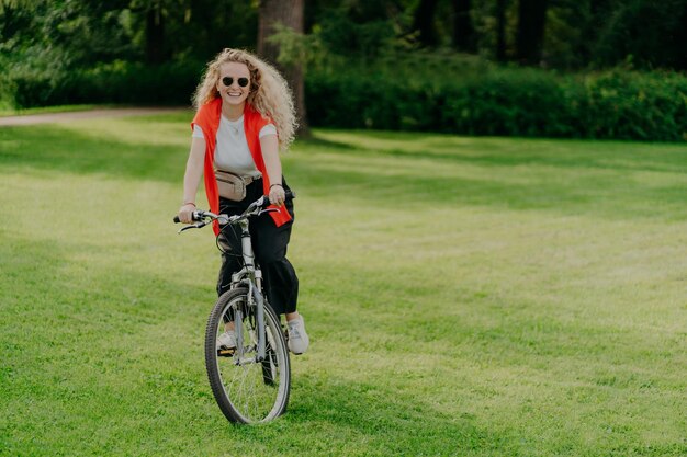 Buiten beeld van vrij vrolijke jonge vrouw rijdt fiets draagt zonnebril vrijetijdskleding poses op groen gazon brengt vrije tijd door in park fietsen in prachtige natuur Activiteit en recreatie concept