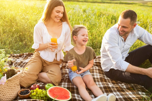 Buiten eten. gelukkige ouders en opgewonden baby rustend en zittend op het gras, sinaasappelsap drinkend, genietend van de dag