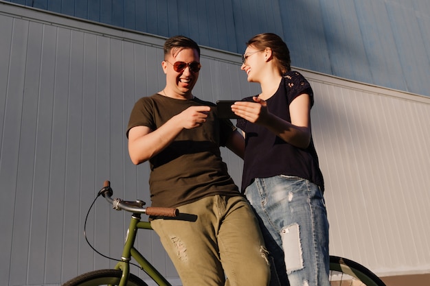 Buiten foto van jonge aantrekkelijke paar, lachen samen tijdens het kijken naar iets grappig op de mobiele telefoon, tijd doorbrengen met plezier, permanent met fiets.