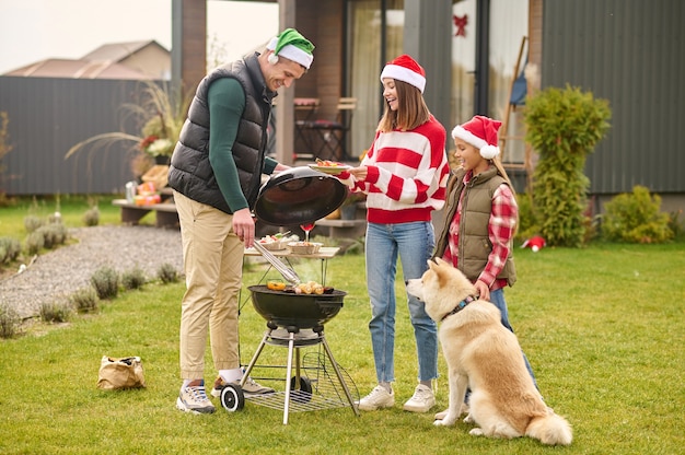 Buiten grillen. Een schattig meisje met een kerstmuts die bij de barbecue staat terwijl haar vader kookt