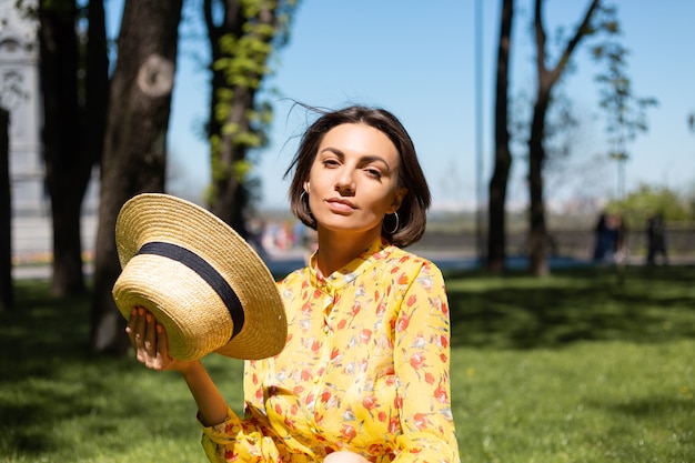 Buiten mode portret van vrouw in gele zomerjurk en hoed zittend op het gras in het park