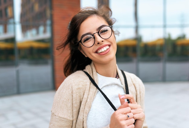 Buiten portret van mooie jonge vrouw student glimlachend breed met brede glimlach met rugzak op de rug dragen transparante bril kijken naar de camera poseren op de straat van de stad