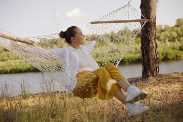 Buiten schot van blanke jonge volwassen vrouw zittend op hangmat aan het water wegkijkend rustend spreidt handen lachend gelukkig met wit overhemd en gele broek