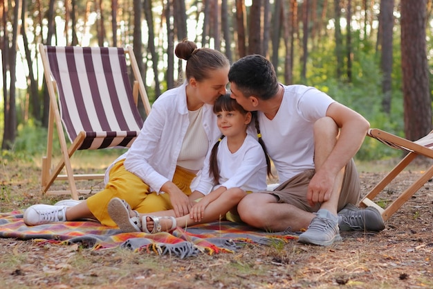 Buiten schot van verliefde paar zittend in het bos met hun kleine schattige dochter ouders kussen hun kind liefde en zachte tijd doorbrengen in hout genieten van frisse lucht