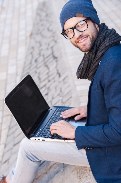 Foto buiten surfen op het net. bovenaanzicht van een knappe jongeman in slimme vrijetijdskleding die op een laptop werkt en glimlacht terwijl hij buiten zit