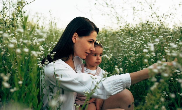 Buiten zijaanzicht beeld van mooie moeder en schattig klein meisje zittend op het groene gras met witte bloemen op het veld verkennen van de natuur Moeder en dochter spelen op een bloemenweide