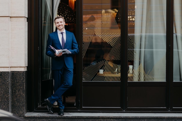 Buitenaanzicht van knappe aantrekkelijke mannelijke directeur of CEO in formeel pak stans gekruiste benen in de buurt van gezellige cafetaria houdt populair dagboek in handen heeft positieve uitdrukking en stoppels Mensen levensstijl