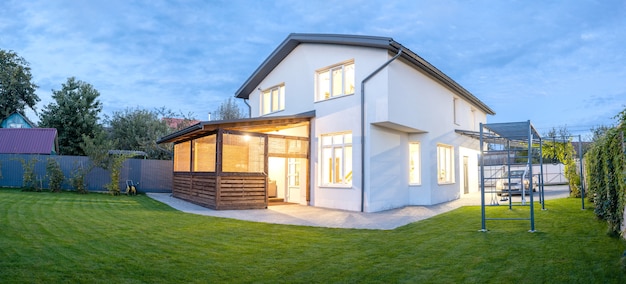 Buitenaanzicht van modern wit huis met binnenplaats, patio, groen grasgazon, tuin en een auto in de avond