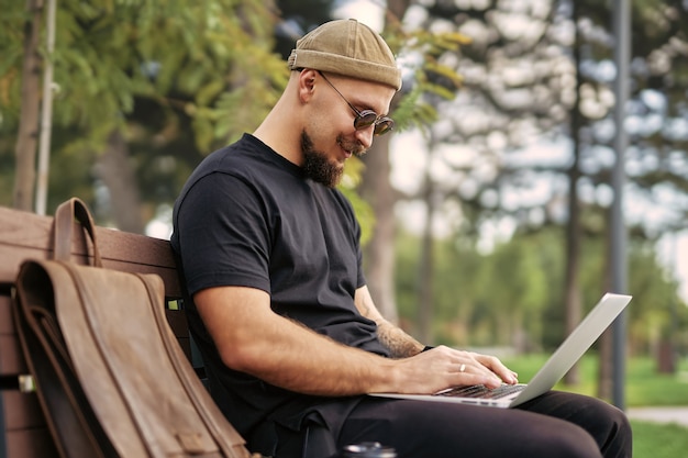 Buitenfoto van positieve millennial in zonnebril die op een bankje zit en op laptop in stadspark werkt