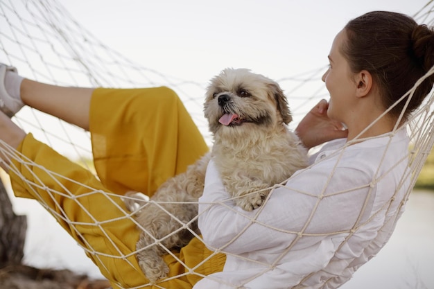 Buitenopname van een donkerharige blanke vrouw met een wit overhemd ligt in een hangmat met Pakingese hond aan de oever van de rivier, ver in de verte kijkend genietend van vrije tijd pratende telefoon