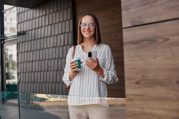 Buitenopname van een kaukasische lachende aantrekkelijke vrouw in elegante kleding die op straat staat in de buurt van een gebouw dat afhaalkoffie drinkt en een mobiele telefoon gebruikt om online te chatten
