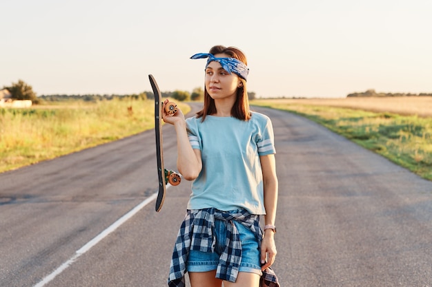 Buitenopname van een mooie donkerharige vrouw die haarband, t-shirt en short draagt, longboard in handen houdt en wegkijkt met doordachte uitdrukking.