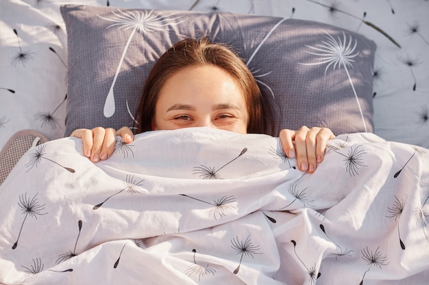 Buitenportret van een donkerharige vrouw die in bed in de open lucht onder zonlicht ligt, haar gezicht onder de deken verbergt, geluk uitdrukt, ontspant en geniet van de natuur.