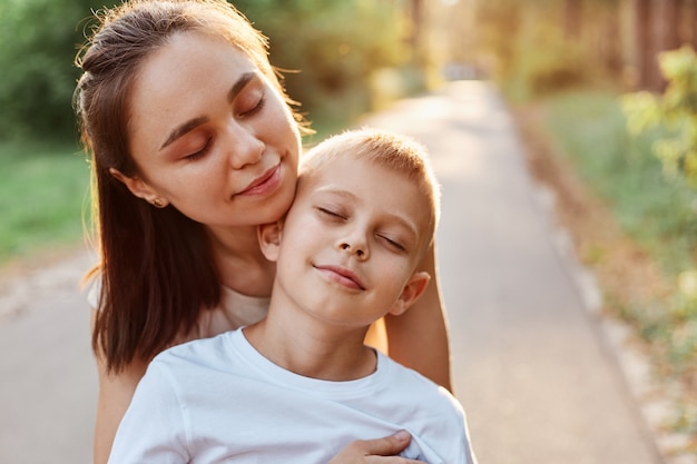 Buitenportret van gelukkige familie, moeder die haar zoon knuffelt, geniet van tijd samen doorbrengen, ogen gesloten houden, wandelen in het zomerpark, moederschap, kindertijd.