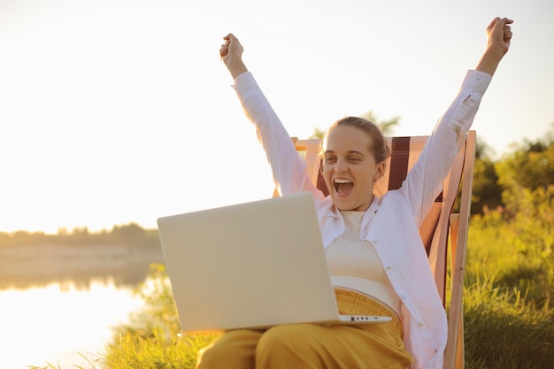 Buitenschot van optimistische opgetogen vrouw met een wit overhemd aan het water in een klapstoel en bezig met laptop met opgeheven armen, blij dat ze in een zeer goed humeur is