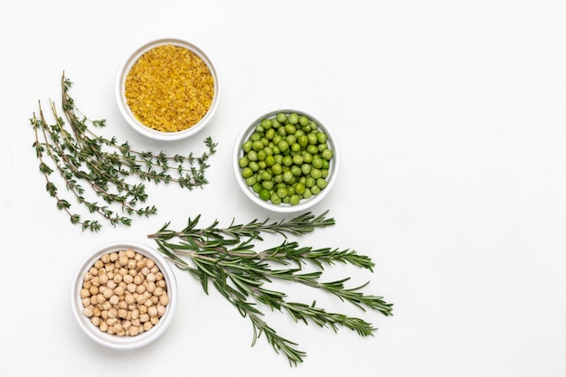 Bulgur, chickpeas and green peas in bowls, sprigs of thyme and rosemary. Copy space. Flat lay. White background