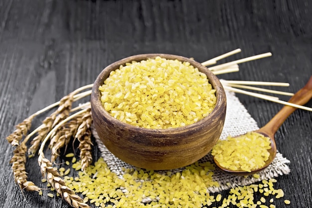 Bulgur groats - steamed grains of wheat - in a clay bowl and a spoon on sacking, ears of wheat on the background of dark wooden board