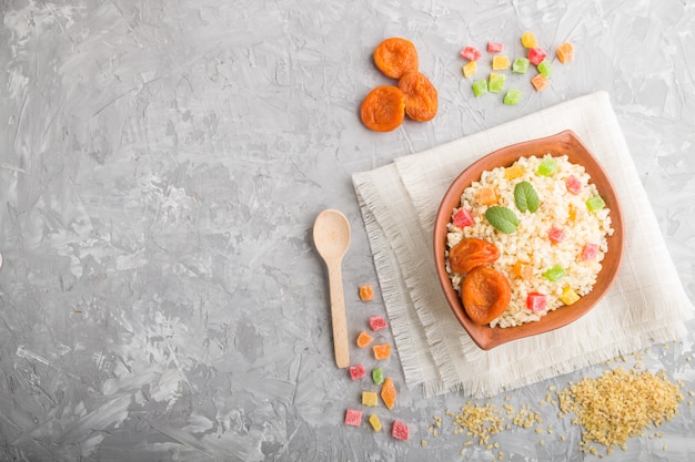 Bulgur porridge with dried apricots and candied fruits in clay bowl on a gray concrete background and linen textile. Top view, copy space.