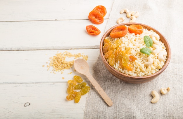 Bulgur porridge with dried apricots,  raisins and cashew in wooden bowl on a white wooden surface and linen textile. Side view, copy space.