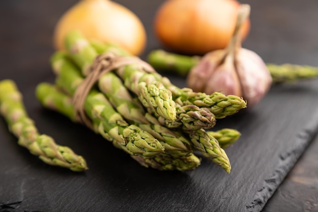 Photo bunch of fresh green asparagus garlic onion on slate board on black concrete background side view close up selective focus