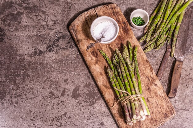 Bunch of fresh green asparagus on vintage wooden chopping board. Delicious ingredients ready for cooking healthy food. Modern hard light, dark shadow. Stone concrete background, top view